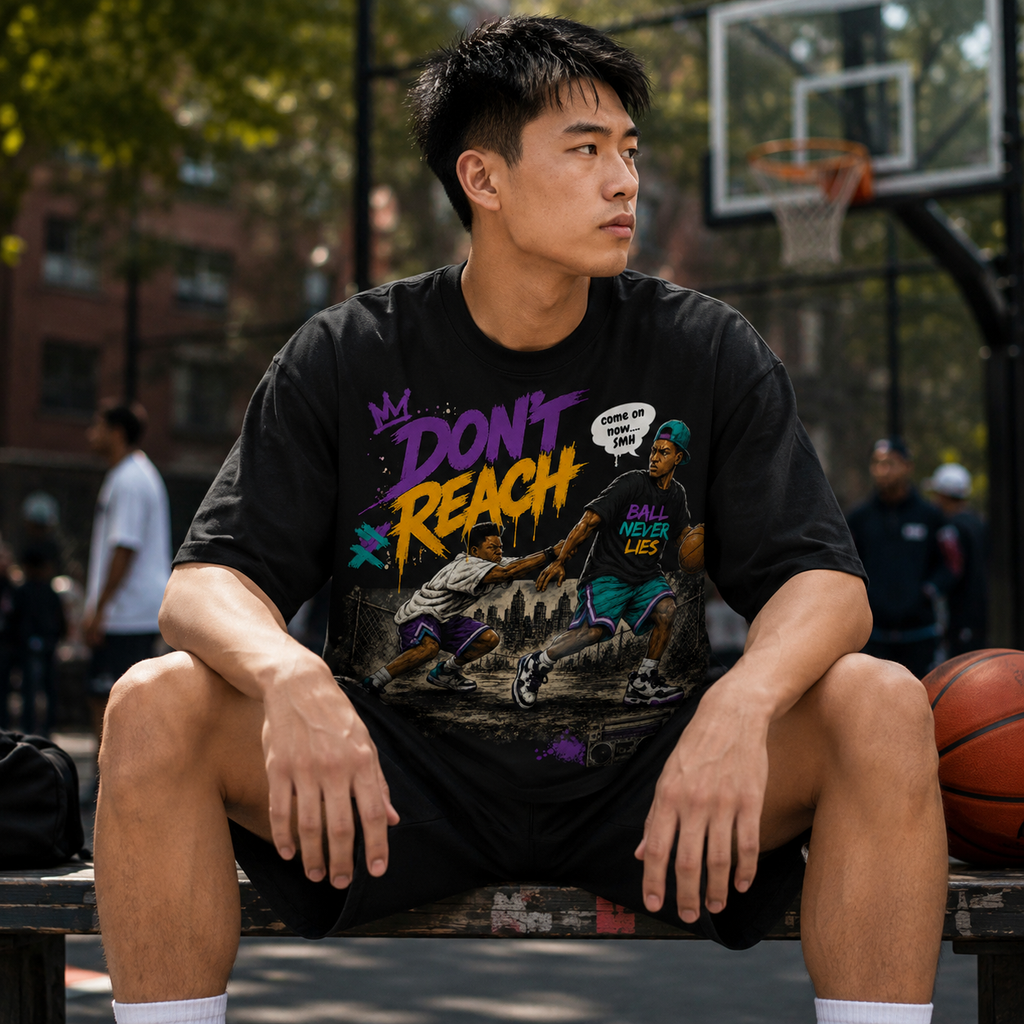 Person wearing a black t-shirt with colorful graphics and text, sitting on a basketball court.