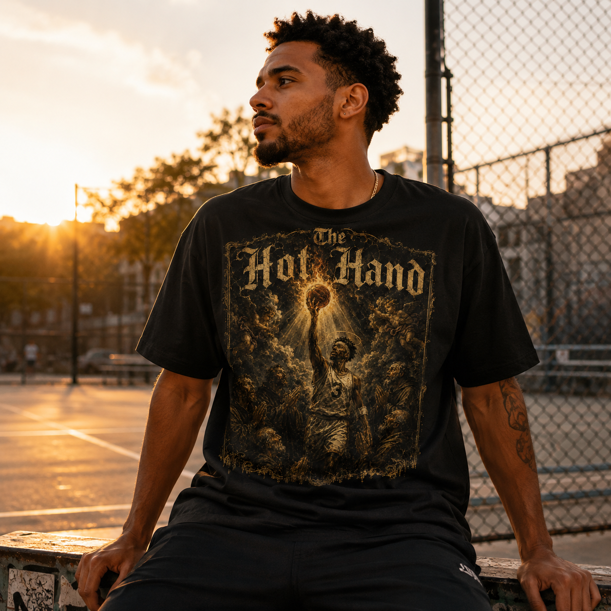 Man wearing a black t-shirt with 'The Hot Hand' design, sitting on a bench outdoors at sunset.