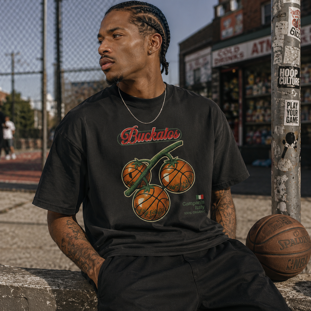 Man wearing a black t-shirt with a graphic design, sitting on a ledge with a basketball, in an urban setting.