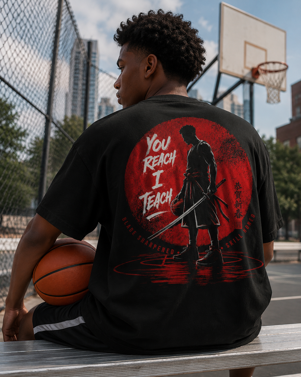 Person wearing a black t-shirt with a graphic design and text, sitting on a basketball court.