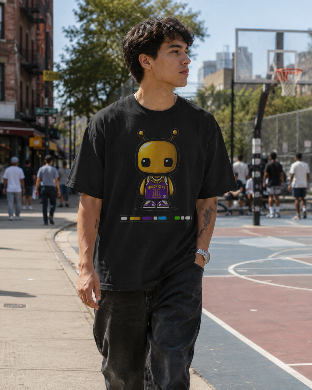 Person wearing a black t-shirt with a graphic design, standing on a sidewalk with a basketball court in the background.