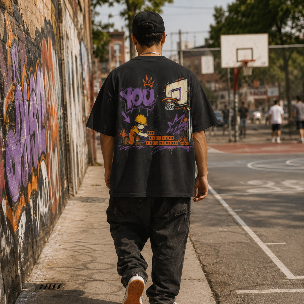Person wearing a black t-shirt with colorful graphics walking on a sidewalk next to a graffiti-covered wall.